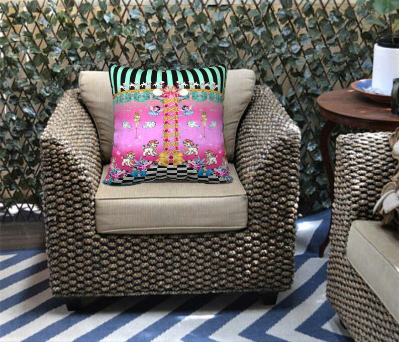 May include: A pink and green patterned throw pillow with a whimsical design featuring a carousel with animals and a checkered floor. The pillow is on a wicker chair with a striped rug.