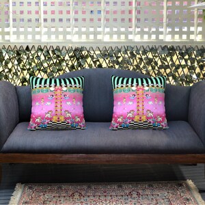 May include: Two pink and black patterned throw pillows with a whimsical design featuring a carousel and other colorful details are placed on a gray sofa with wooden arms. The sofa is in front of a white lattice fence with green ivy.
