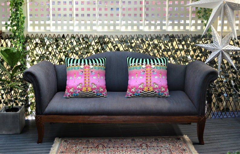 May include: Two pink and black patterned throw pillows with a whimsical design featuring a carousel and other colorful details are placed on a gray sofa with wooden arms. The sofa is in front of a white lattice fence with green ivy.