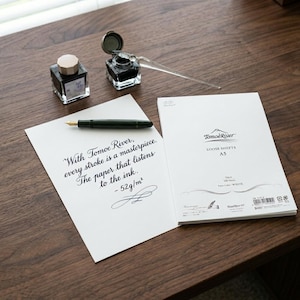May include: A still-life composition featuring stationery items on a wooden surface. Includes a fountain pen, ink bottles, a glass dip pen, and a stack of white paper with the text "With Tomoe River, every stroke is a masterpiece."