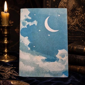 Op de afbeelding: Een blauw notitieboek met een hemels ontwerp met een halve maan, sterren en wolken. De omslag heeft een aquareleffect. Een brandende kaars en antieke boeken staan op de achtergrond en creëren een vintage esthetiek.
