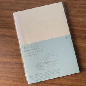 May include: A rectangular notebook with cream-colored pages enclosed in a clear plastic wrapper. A light blue paper band wraps around the lower portion, featuring a line drawing of a hand writing on a grid and text including "Designed for Writer's Comfort" and "MD Notebook A5 Gridded."
