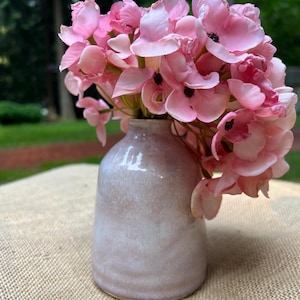 May include: A small, rounded ceramic vase with a smooth, pale pink and white glazed finish. The vase is filled with a dense cluster of artificial pink flowers, each featuring delicate, layered petals and small, dark centers at their core.