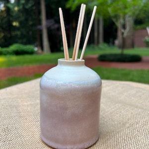 May include: A cylindrical ceramic diffuser vessel with a smooth, gradient finish transitioning from a soft, pale cream at the neck to a muted, dusty mauve-pink at the base. Four thin, light-colored wooden reeds extend vertically from the opening.
