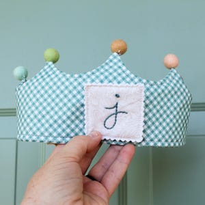 Può includere: Una corona di stoffa fatta a mano con un motivo a quadri verde e bianco. La corona ha una piccola toppa quadrata con la lettera "j" ricamata. La corona è sormontata da pompon colorati.