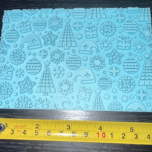 May include: A light blue rectangular silicone texture mat featuring a repeating embossed pattern of Christmas trees, snowflakes, gift boxes, ornaments, and Santa hats, positioned above a yellow measuring tape showing a width of 15 cm.