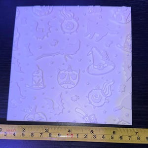 May include: A square white embossing plate featuring a repeating pattern of Halloween icons including a witch hat, a jack-o'-lantern, a cat, a cauldron, and bats. A yellow measuring tape is positioned at the bottom, showing a width of 17.8 cm.