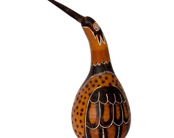 Op de afbeelding: Een kalebassculptuur in de vorm van een vogel met een lange, puntige snavel. Het oppervlak is beige met donkerbruine geschilderde details, waaronder een reeks stippen op het lichaam, gestileerde vleugelveren en een cirkelvormig oogpatroon op de kop.