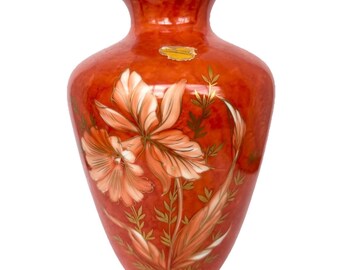 Op de afbeelding: Een decoratieve oranje vaas met een gouden rand en bloemenmotief. De vaas heeft grote, lichtoranje bloemen met gouden accenten en groene bladeren. Aan de zijkant is een kleine gouden sticker bevestigd.
