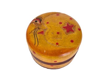 Op de afbeelding: Een ronde houten doos met een gele afwerking, voorzien van een geschilderd maangezicht met zwarte lijndetails, een rode ster en kleine rode stippen op het deksel en de zijkanten. Een donkerbruine band loopt horizontaal rond de onderkant.