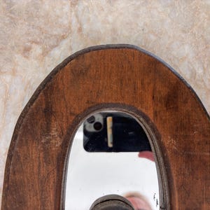 May include: A curved, dark brown wooden frame surrounding a central mirror. The wood displays a visible grain and natural texture. The mirror surface reflects a black smartphone and a portion of a person's hand, showing a clear, reflective finish.