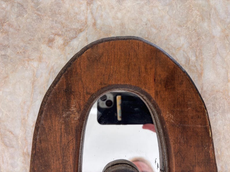 May include: A curved, dark brown wooden frame surrounding a central mirror. The wood displays a visible grain and natural texture. The mirror surface reflects a black smartphone and a portion of a person's hand, showing a clear, reflective finish.