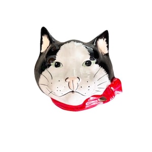 May include: A ceramic wall plaque shaped like a cat head with black and white markings, green eyes, and a pink nose. The cat is wearing a bright red neckerchief tied in a bow on the right side. The surface is glossy with painted whiskers.
