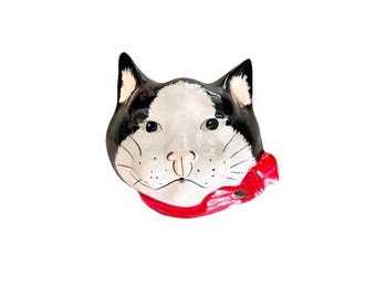 Op de afbeelding: Een keramieken wandbord in de vorm van een kattenkop met zwart-witte markeringen, groene ogen en een roze neus. De kat draagt een felrode halsdoek die aan de rechterkant in een strik is geknoopt. Het oppervlak is glanzend met geschilderde snorharen.