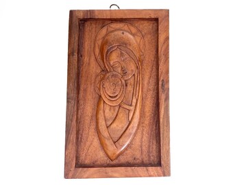 Op de afbeelding: Een rechthoekige bruine houten wandplaat met een handgesneden reliëf van een figuur die een kind vasthoudt. Beide figuren hebben cirkelvormige halo's rond hun hoofd gegraveerd. Het stuk heeft een gladde afwerking en een metalen haakje.