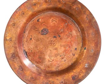 Op de afbeelding: Een ronde koperen schaal met een verweerde, geoxideerde patina en een gevlekt oranje en bruin oppervlak. De brede rand is versierd met een herhalend patroon van kleine, gestempelde ruitvormige motieven, terwijl het midden vage, geëtste symbolen toont.