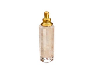 Op de afbeelding: Een kleine, cilindervormige fles van helder glas met een gefacetteerd, getextureerd oppervlak en een gladde, afgeronde dop van goudkleurig metaal. De fles heeft een verticaal, veelzijdig ontwerp en de dop heeft een getrapte vorm.