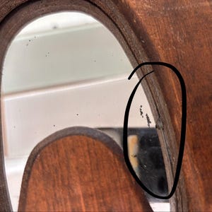 May include: A close-up view of a wooden mirror frame with a dark brown finish. The reflective glass surface shows small black spots and imperfections near the edge, which is highlighted by a hand-drawn black circle on the image.