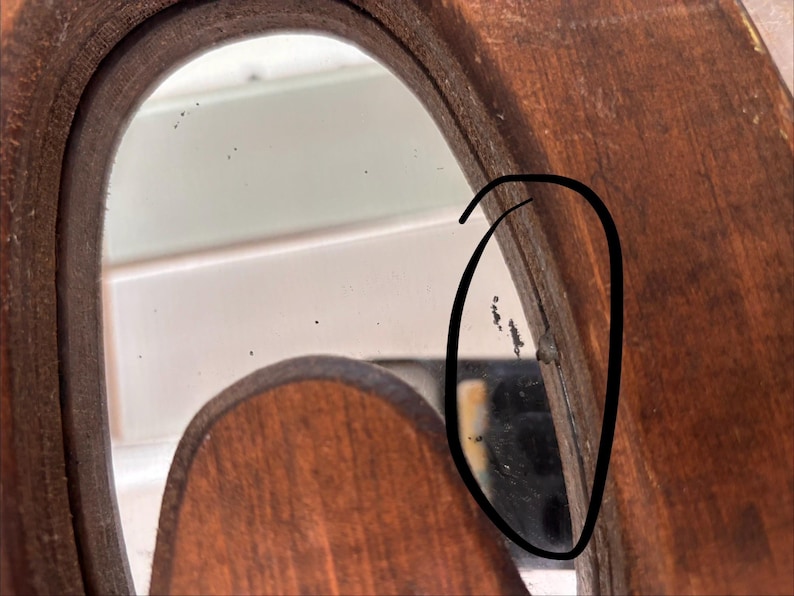 May include: A close-up view of a wooden mirror frame with a dark brown finish. The reflective glass surface shows small black spots and imperfections near the edge, which is highlighted by a hand-drawn black circle on the image.