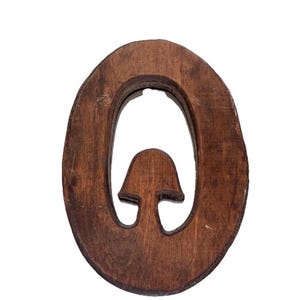 May include: A dark brown wooden cutout in the shape of the letter O, featuring a small, centered mushroom-like silhouette carved within the open space of the letter. The wood grain is visible throughout the surface of the letter-shaped object.
