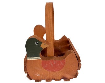 Op de afbeelding: Een houten mand met een eendvormig zijpaneel met een donkergroene kop, een beige snavel en een rood vleugeldetail. De mand heeft een hoog, gebogen houten handvat en een natuurlijke houtnerfafwerking, opgebouwd uit meerdere houten latten.