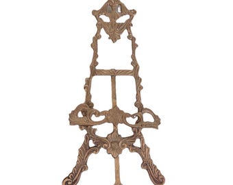 Op de afbeelding: Een hoge, sierlijke koperen ezelstandaard met ingewikkeld krulwerk en bloemmotieven. De structuur bevat een centrale verticale steunstang, een horizontale richel voor het uitstallen van items en een decoratieve puntige top met metalen afwerking.