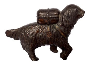 Op de afbeelding: Een bronzen hondenbeeldje met een rugzak. De hond wordt afgebeeld in een loopstand, met gedetailleerde vachttextuur en een riem die de rugzak vastzet. De rugzak is rechthoekig en lijkt van leer te zijn.