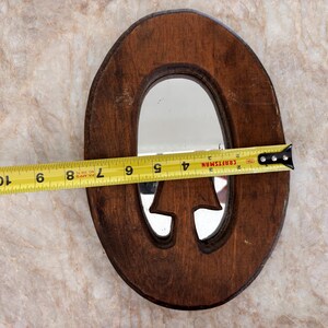 May include: An oval-shaped wooden mirror with a dark brown finish and a cutout mushroom-shaped detail at the bottom. A yellow Craftsman measuring tape is positioned across the center, showing a width of approximately 7.75 inches for the mirror frame.