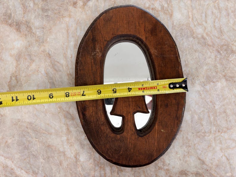 May include: An oval-shaped wooden mirror with a dark brown finish and a cutout mushroom-shaped detail at the bottom. A yellow Craftsman measuring tape is positioned across the center, showing a width of approximately 7.75 inches for the mirror frame.
