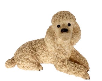 Op de afbeelding: Een beige poedelfiguur in liggende positie. Het beeldje is voorzien van gedetailleerde, gestructureerde krullende vacht over het hele lichaam, met donkere, ronde ogen en een kleine zwarte neus. De hond rust op zijn voorpoten met de kop omhoog.