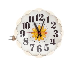 May include: A round, scalloped-edge wall clock with a cream-colored frame and a white face. The clock features large black numerals, a central yellow flower graphic, black hour and minute hands, and a thin red second hand. A power cord is attached.
