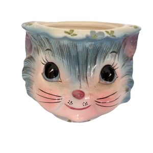 May include: A ceramic planter shaped like a cat's face. The planter has a light blue and pink color scheme, with black eyes, pink cheeks, and a small pink nose. The top rim is decorated with small green leaves and white flowers.