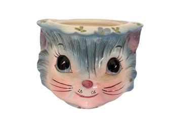 Op de afbeelding: Een keramische plantenbak in de vorm van een kattengezicht. De plantenbak heeft een lichtblauw en roze kleurenschema, met zwarte ogen, roze wangen en een kleine roze neus. De bovenrand is versierd met kleine groene blaadjes en witte bloemen.