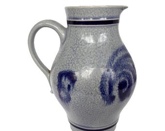 Op de afbeelding: Een grijze keramische kan met een handvat en tuit. De kan heeft een getextureerd oppervlak en is versierd met donkerblauwe bloemmotieven en banden. Deze kan in vintage-stijl is geschikt voor het serveren van dranken.