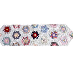 May include: A white, quilted table runner with a repeating floral pattern. The flowers are in various colors, including red, pink, blue, and yellow. The runner is approximately 60 inches long.