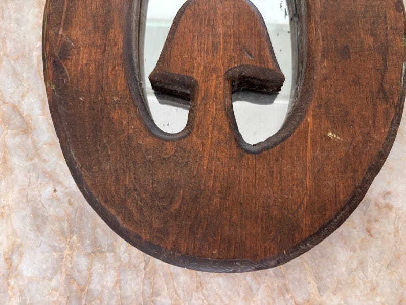 May include: A circular wooden mirror frame with a dark brown finish and a visible wood grain texture. The frame features a central cutout in the shape of a mushroom, with the reflective mirror surface visible through the opening of the cutout.