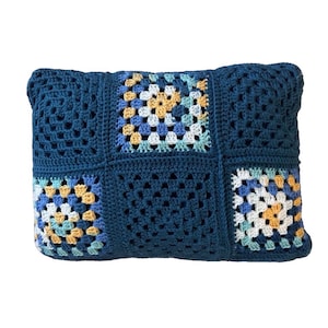 May include: A rectangular crochet pillow featuring a patchwork design of solid dark blue squares and multi-colored granny squares in shades of yellow, light blue, white, and teal. The textile construction displays a repeating yarn stitch pattern.