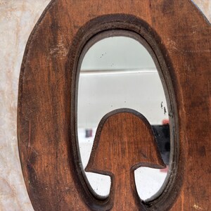 May include: A brown wooden mirror frame with a natural, weathered texture and an oval-shaped reflective glass center. The frame features a carved, mushroom-shaped wooden element extending into the lower portion of the mirror's reflective surface.