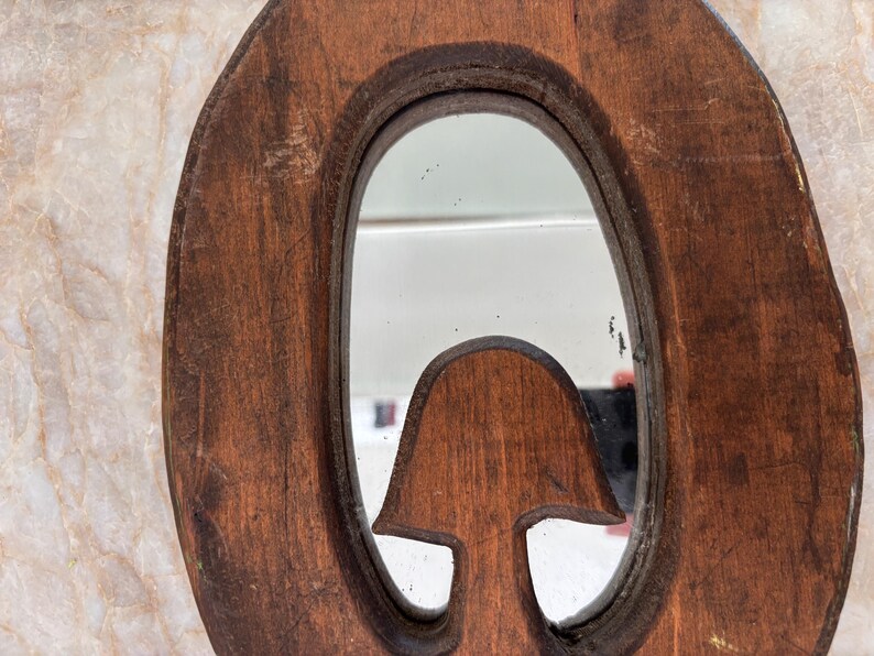 May include: A brown wooden mirror frame with a natural, weathered texture and an oval-shaped reflective glass center. The frame features a carved, mushroom-shaped wooden element extending into the lower portion of the mirror's reflective surface.