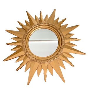 May include: A circular wall mirror featuring a gold-colored frame shaped like a sunburst. The frame consists of alternating straight and wavy rays radiating from a central circular glass pane, which is surrounded by a decorative beaded border.