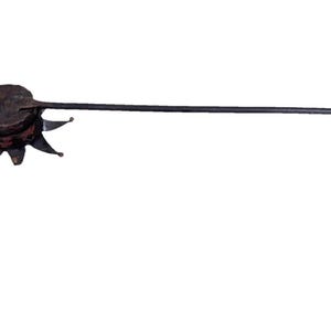 May include: A dark brown, circular metal object with a textured surface and a star-shaped base featuring pointed tips. A long, thin, straight metal handle extends from the side, ending in a small, rounded knob at the far tip of the tool.