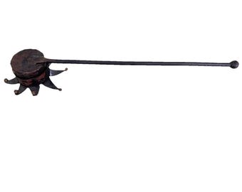 Op de afbeelding: Een donkerbruin, cirkelvormig metalen object met een getextureerd oppervlak en een stervormige basis met puntige uiteinden. Een lange, dunne, rechte metalen steel steekt uit de zijkant en eindigt in een kleine, ronde knop aan het uiteinde.