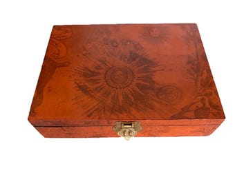 Op de afbeelding: Een rechthoekige oranje houten doos met een koperen sluiting aan de voorzijde. Het deksel is versierd met een donkerbruine print van een zon met een gezicht, hemelse elementen en botanische patronen over het gehele oppervlak van de doos.