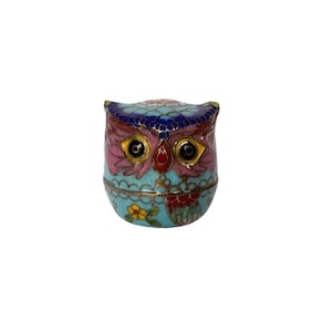 May include: A small, decorative owl-shaped trinket box. The box is primarily turquoise with pink, red, and gold accents. The owl's eyes are black with gold rims. The top of the owl's head is dark blue.