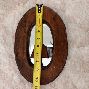 May include: An oval-shaped wooden mirror with a dark brown finish and a carved mushroom detail at the bottom. A yellow Craftsman measuring tape is placed vertically across the center, showing a height of approximately 10.5 inches.