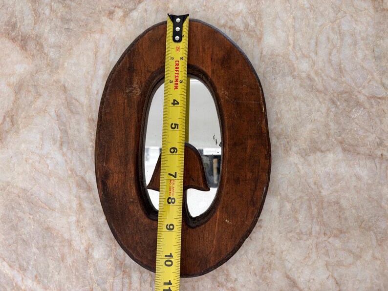 May include: An oval-shaped wooden mirror with a dark brown finish and a carved mushroom detail at the bottom. A yellow Craftsman measuring tape is placed vertically across the center, showing a height of approximately 10.5 inches.