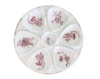 Op de afbeelding: Een witte keramische serveerschaal met zes hartvormige vakjes gerangschikt rond een centrale ronde kom. Elk gedeelte is versierd met een gedetailleerde bordeauxrode bloemenillustratie. De schaal heeft een gestructureerd, rasterachtig oppervlak.
