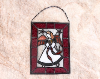 Op de afbeelding: Een rechthoekig glas-in-loodpaneel met een centrale figuur met oranje vleugels en een witte jurk, omringd door een donkerrode rand. Het werk is gevat in zwart metaal en voorzien van een dunne zwarte ketting om het op te hangen.