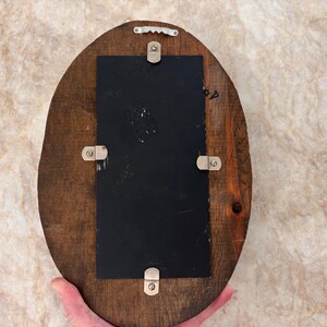 May include: An oval-shaped wooden plaque featuring a dark brown finish with a visible wood grain. A rectangular black panel is centered on the wood, secured by four silver-toned metal clips. A silver-toned sawtooth hanger is attached at the top.