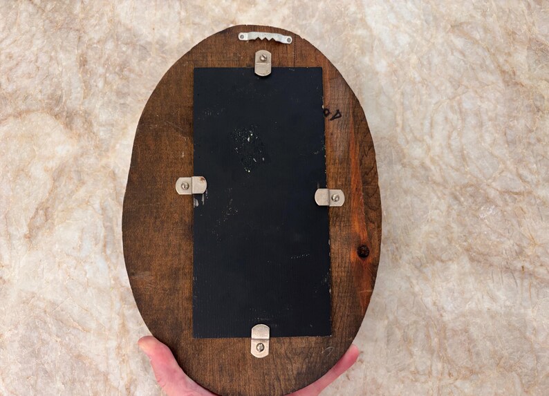 May include: An oval-shaped wooden plaque featuring a dark brown finish with a visible wood grain. A rectangular black panel is centered on the wood, secured by four silver-toned metal clips. A silver-toned sawtooth hanger is attached at the top.